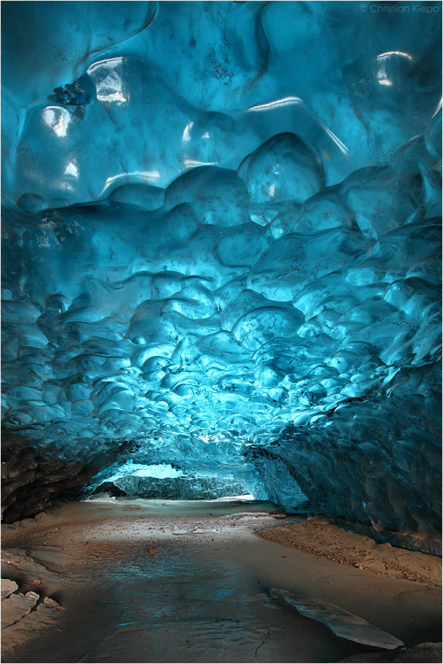 
This up to 1000 years old snow has metamorphosed into highly pressurized  glacier ice that contains almost no air bubbles. Thus it absorbs the  visible light despite the scattered shortest blue fraction, giving it  its distinct deep blue waved appearance. This cavity in the glacier ice  formed as a result of a glacial mill, or moulin.
Rain and meltwater on  the glacier surface is channelled into streams that enter the glacier at  crevices. The waterfall melts a hole into the glacier while the ponded  water drains towards lower elevations by forming long ice caves with an  outlet at the terminus of the glacier. The fine grained sediments in the  water along with wind blown sediments cause the frozen meltwater stream  to appear in a muddy colour while the top of the cave exhibits the deep  blue colour.
Due to the fast movement of the glacier of about 1 m per  day over uneven terrain this ice cave cracked up at its end into a deep  vertical crevice, called cerrac. This causes the indirect daylight to  enter the ice cave from both ends resulting in homogeneous lighting of  the ice tunnel.

