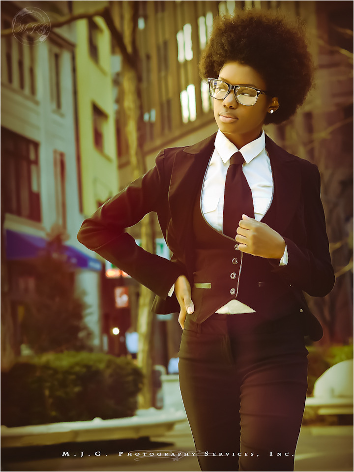 Person from knees up; rocks a tie and fitted suit, vest, colared shirt, RayBans, natural kinky hair,  poses and glances off-screen. Dusk city scape.
