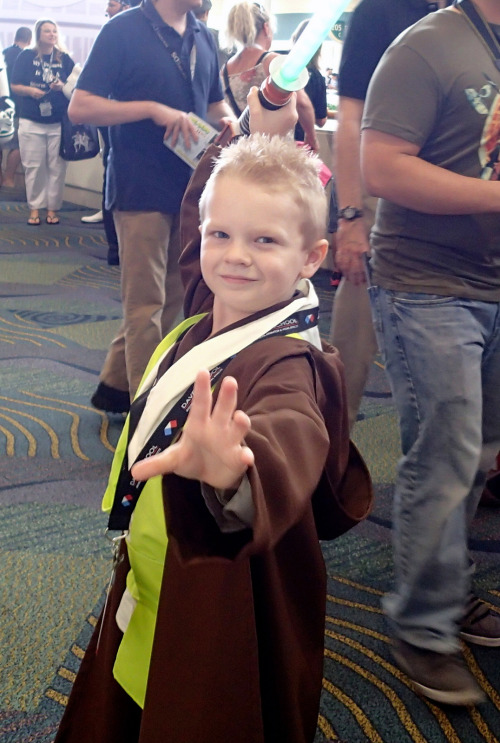 A blonde young man dressed as a Jedi holding his lightsaber arced above his head in a classic Jedi pose. 